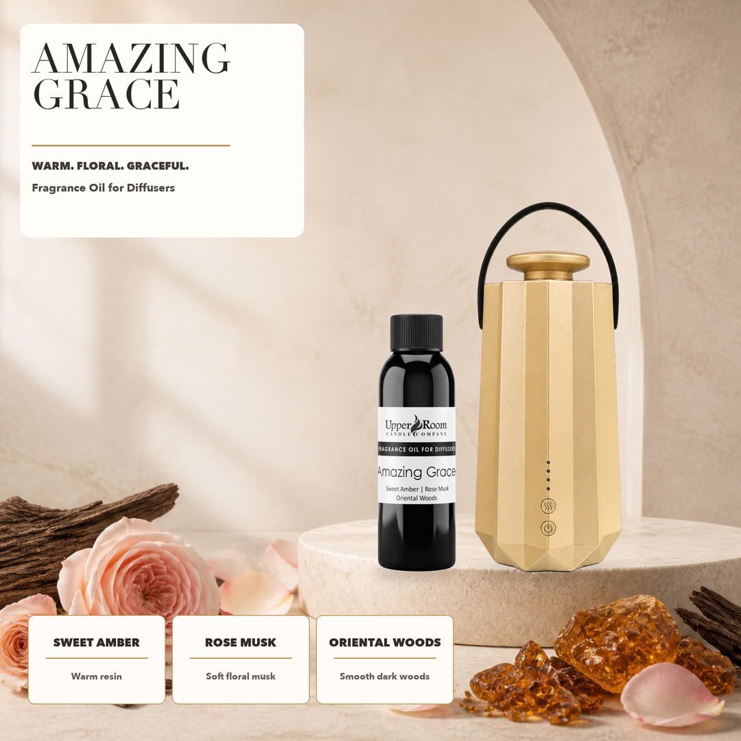 A fragrance oil diffuser set labeled Amazing Grace, featuring a faceted gold diffuser with a black handle, a small black bottle of fragrance oil, and decorative amber pieces and roses on a beige stone surface. Text cards describe the scent notes: Sweet Amber, Rose Musk, and Oriental Woods.