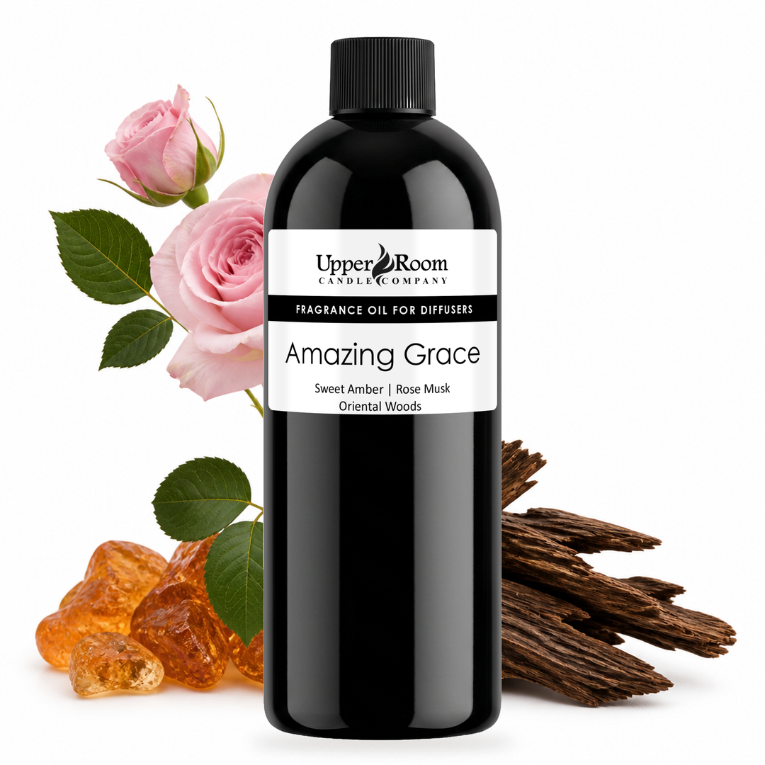 A black bottle of Upper Room Candle Company fragrance oil labeled 'Amazing Grace' stands centered on a white background, styled with pink roses, green leaves, amber pieces, and brown wood slices.