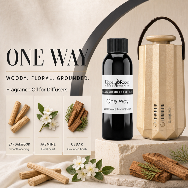 A bottle of Upper Room 'One Way' fragrance oil stands beside a beige diffuser on a stone pedestal, with scent notes for sandalwood, jasmine, and cedar shown in a graphic panel. The scene is styled with wood chips, white flowers, and greenery on a warm beige background.