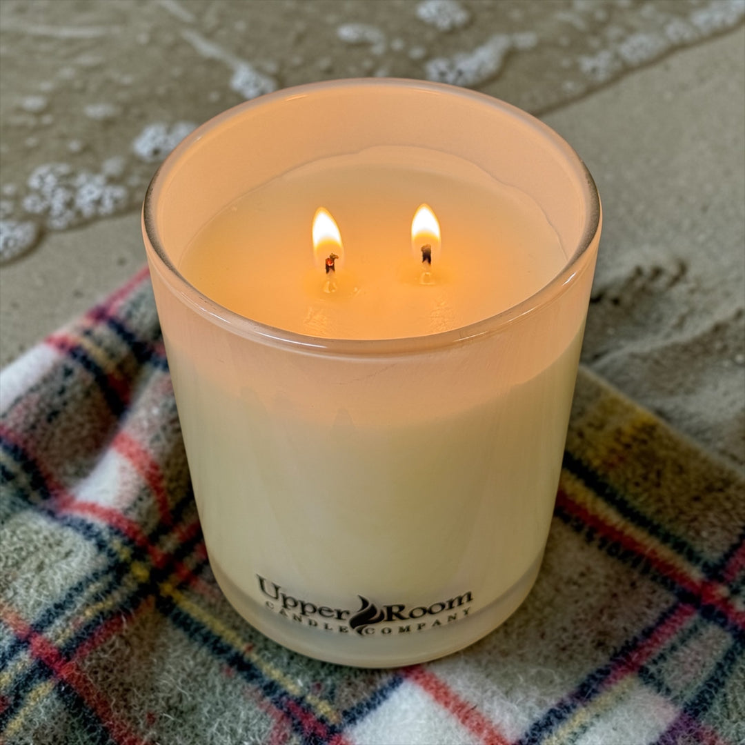 Cashmere Coast Candle, double wick gold jar on a cashmere blanket on the beach near the water line, a wave coming in close, upper room candle company