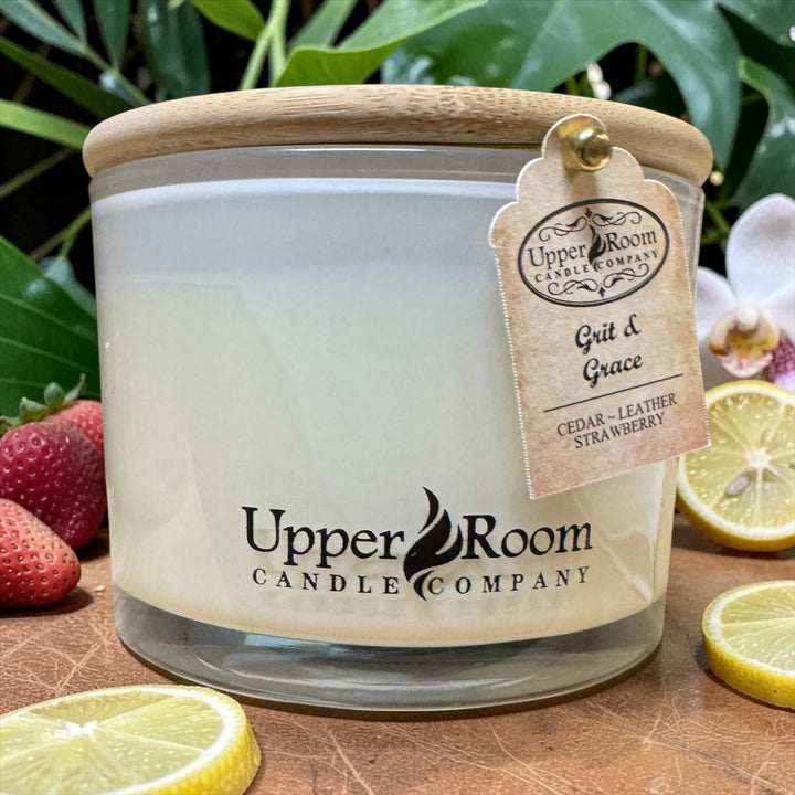 Grit & Grace Candle, triple wick candle, white jar, in front of strawberries and lemons on a leather table, in front of large leaved plants, upper room candle company